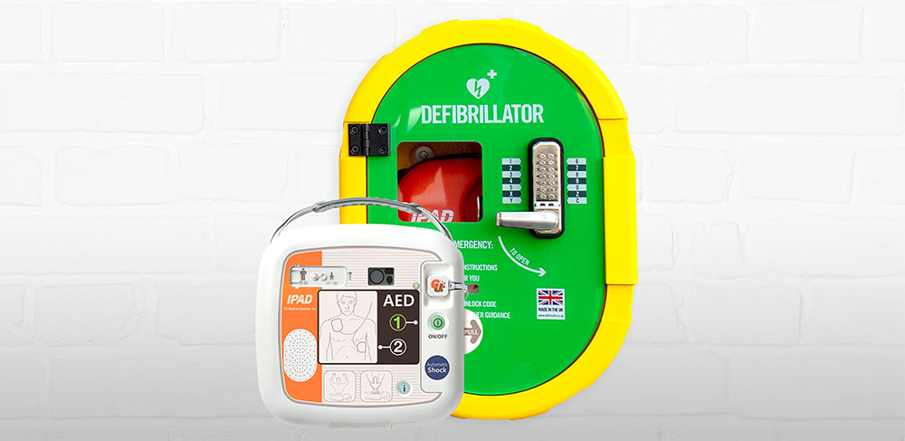A close-up image of an AED (automated external defibrillator) device alongside a bright yellow and green defibrillator case mounted on a white brick wall. The case has a combination lock and instructions for use.