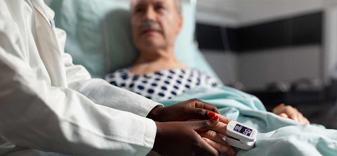 Friendly doctor reassuring sick old patient holding hands using oximeter