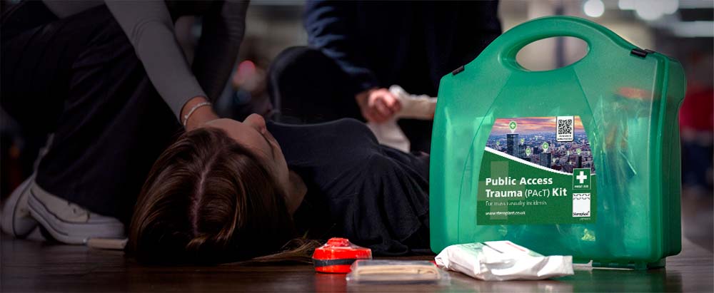Green Public Access Trauma (PAcT) Kit prominently displayed in the foreground, with emergency medical supplies scattered beside it. In the background, a person lies on the floor while two others attend to them, suggesting a medical emergency. The setting is indoors, with a blurred background emphasising the kit and responders.