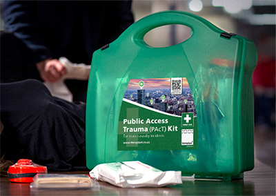Close-up view of a green Public Access Trauma (PAcT) Kit case with its label visible, placed on the floor alongside first aid supplies. In the background, part of a person is seen attending to someone lying down, indicating an emergency response scenario. The focus is on the kit, with the blurred background suggesting an indoor setting.