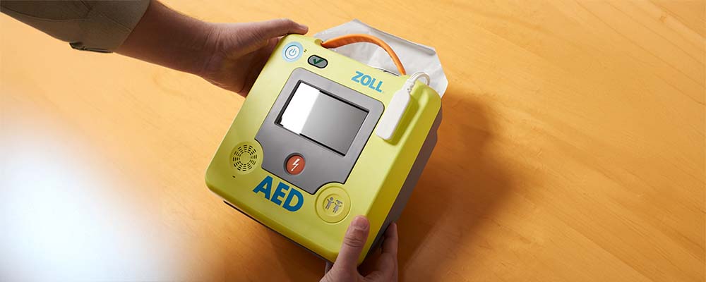 Hands holding a ZOLL AED 3 device, displaying its touchscreen and design.