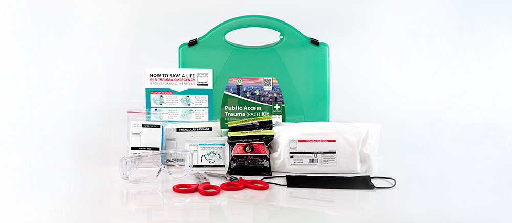 Public Access Trauma (PAcT) Kit displayed with its contents spread out. The green carrying case is positioned at the back, with various emergency supplies in front, including safety glasses, red trauma scissors, a tourniquet, bandages, a face mask, instructional leaflets, and other medical items. The kit is organised to provide essential tools for trauma emergencies.