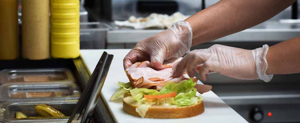 Restaurant kitchen employee in gloves placing deli meat on bread to make sandwich order for customer