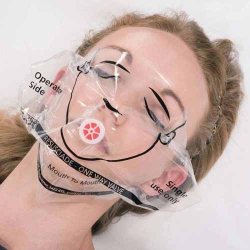Person lying down with a Resusciade face shield placed over their face, showing the shield's one-way valve positioned over the mouth. The shield is transparent, with printed facial guidelines and labelled instructions such as 'Operator Side' and 'Single Use Only' for correct positioning.