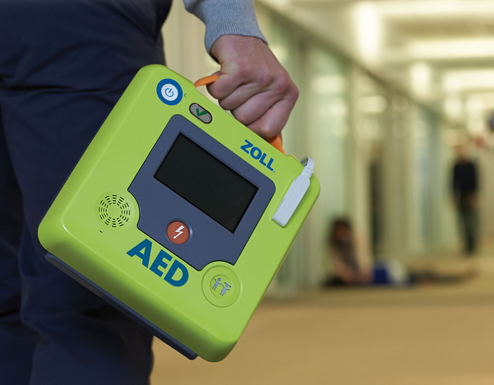 A person holding a bright green Zoll AED (Automated External Defibrillator) by its handle, walking through a hallway, with a blurred figure lying on the ground in the background