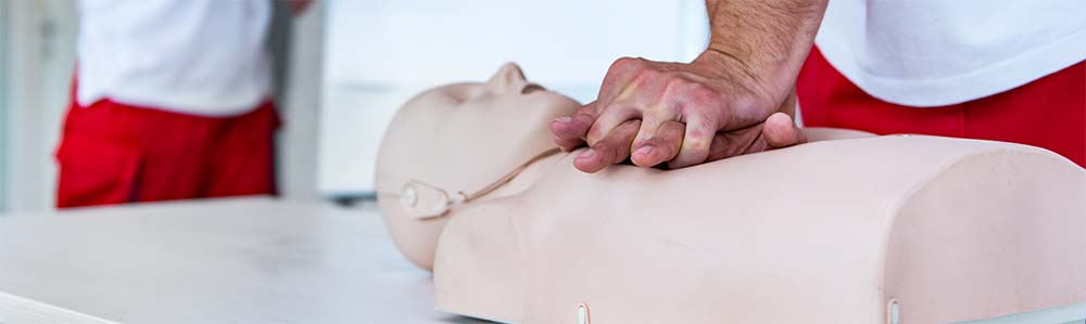 A person practising chest compressions on a CPR training manikin. The person is wearing a red outfit, and the manikin is laid out on a flat surface, suggesting a training environment.