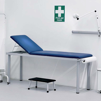 A medical room with a blue examination couch, a small step stool, and a wall-mounted first aid sign above the bed.