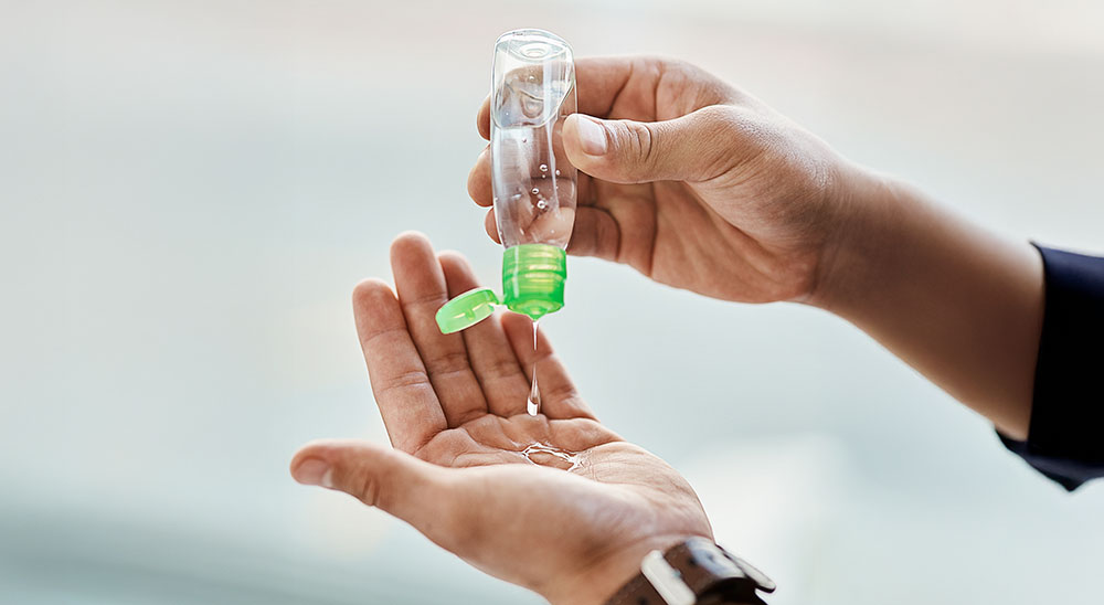 A close-up of a person squeezing hand sanitiser from a small transparent bottle with a green flip cap onto their palm. The person's other hand, wearing a brown leather strap watch, is held out to receive the sanitiser.