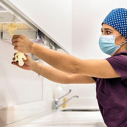 A healthcare worker in a mask and scrubs, pulling medical gloves from a wall dispenser in a clean medical environment