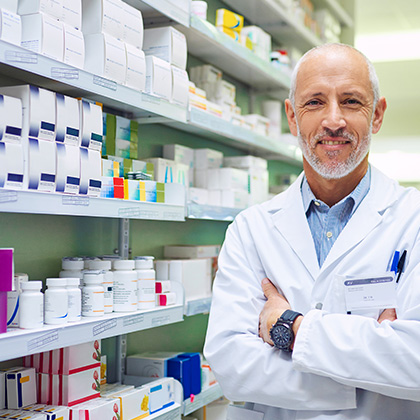 A smiling pharmacist standing confidently with arms crossed in front of well-stocked shelves of medications in a pharmacy